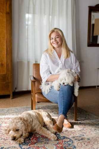 Stephanie Illich sitzt entspannt mit einem weißen kleinen Hund auf dem Schoß, während ein Goldendoodle zu ihren Füßen liegt